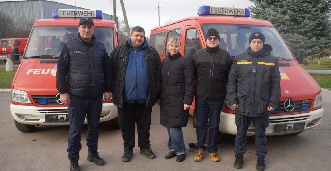 Foto: e 5 Menschen stehen vor den beiden Feuerwehrfahrzeugen.
