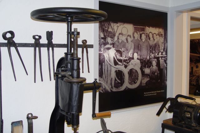 links ein Standbohrmaschine mit Handbetrieb (um 1890), rechts ein Elektromotor. Im Hintergrund die "Mechanische Werkstatt Theodor Müller" in Bernhausen, Foto um 1930.